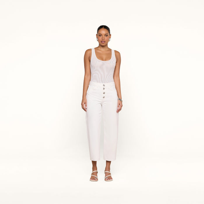 A woman stands facing forward, styled in the Kalea Mesh Bodysuit beneath high-waisted white pants with buttons and white sandals, set against a plain white background.