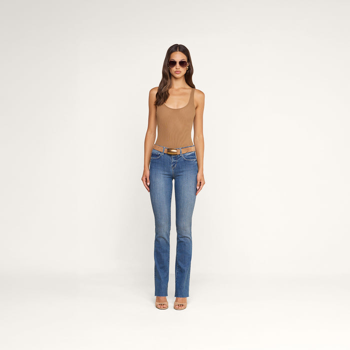 A woman stands against a plain white background wearing sunglasses, blue jeans with a belt, heeled sandals, and the Kalea Mesh Bodysuit in tan. She faces forward with her arms relaxed at her sides.