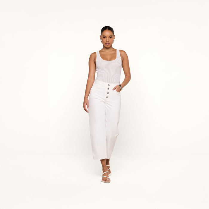 Wearing the Kalea Mesh Bodysuit styled as a white tank top, paired with high-waisted white pants with buttons and white strappy sandals, a woman stands against a plain white background, confidently looking at the camera with one hand in her pocket.