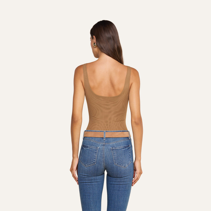 A woman with long brown hair, wearing the sleeveless Kalea Mesh Bodysuit in tan paired with blue jeans, stands with her back to the camera against a plain light background.
