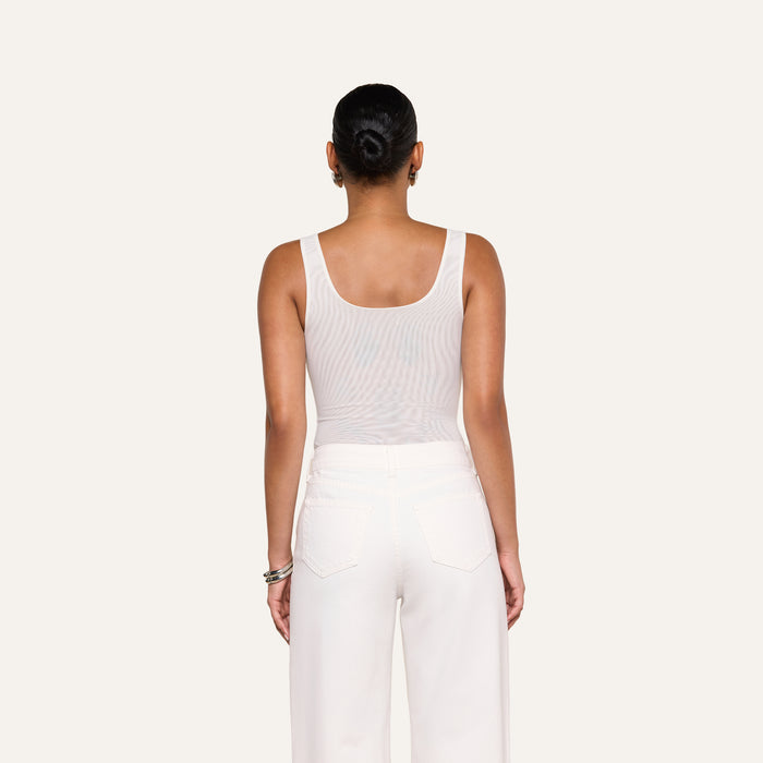 A person stands facing away from the camera, wearing the Kalea Mesh Bodysuit in white and white pants against a plain light background.