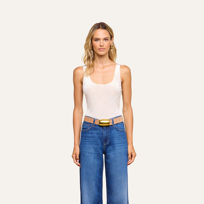 A woman with wavy blonde hair wears a sleeveless white Kalea Velvet Bodysuit, blue jeans, and a tan belt with a gold buckle, standing against a plain light background.