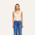 A woman with wavy blonde hair wears a sleeveless white Kalea Velvet Bodysuit, blue jeans, and a tan belt with a gold buckle, standing against a plain light background.