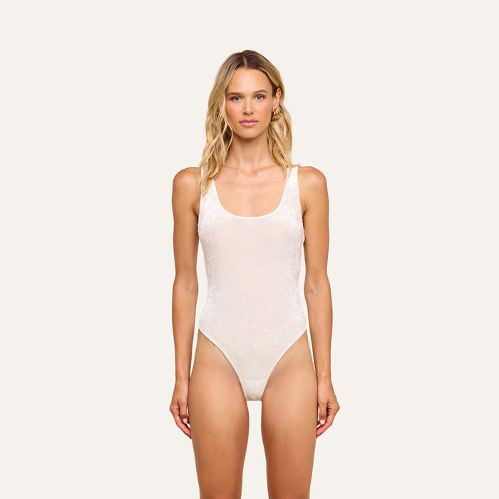 A woman with wavy blonde hair faces forward, wearing the Kalea Velvet Bodysuit in ivory as a lightweight layer against a plain, light background.