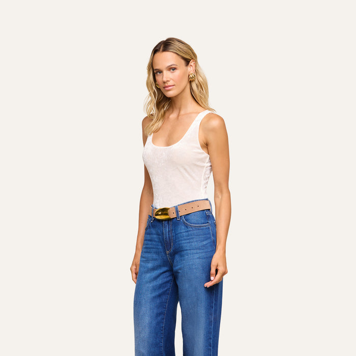 A woman with long blonde hair wears the Kalea Velvet Bodysuit as a sleeveless white top, paired with blue high-waisted jeans and a tan belt, standing against a plain light background.