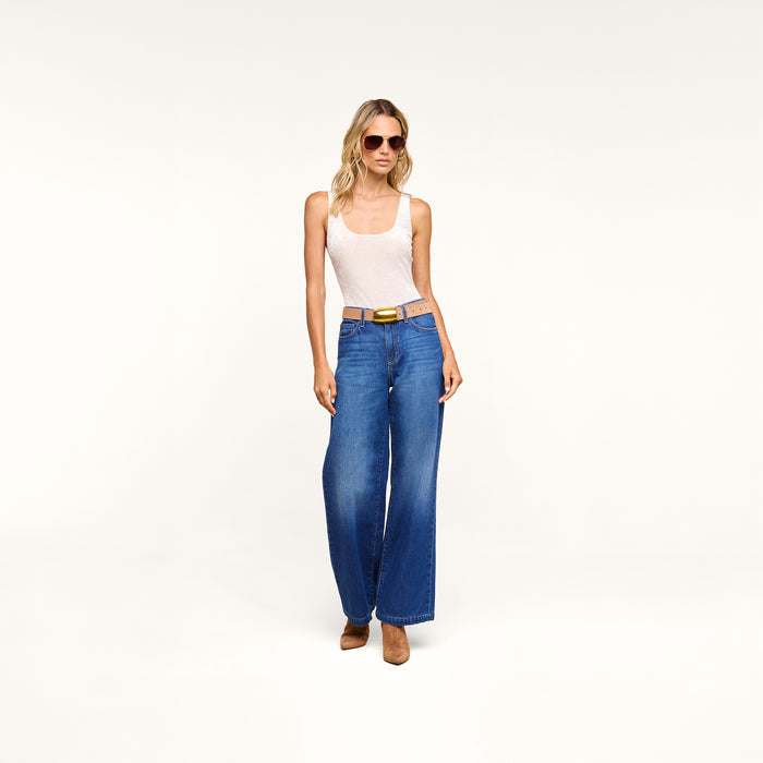 Woman wearing sunglasses, a Kalea Velvet Bodysuit styled as a white tank top, blue wide-leg jeans with a belt, and brown heels stands confidently against a plain white background.