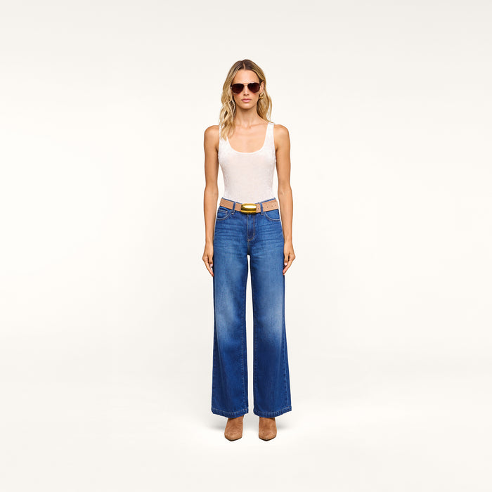 Woman wearing sunglasses, the Kalea Velvet Bodysuit styled as a lightweight layer, blue wide-leg jeans, brown belt, and brown shoes stands facing forward against a plain white background.