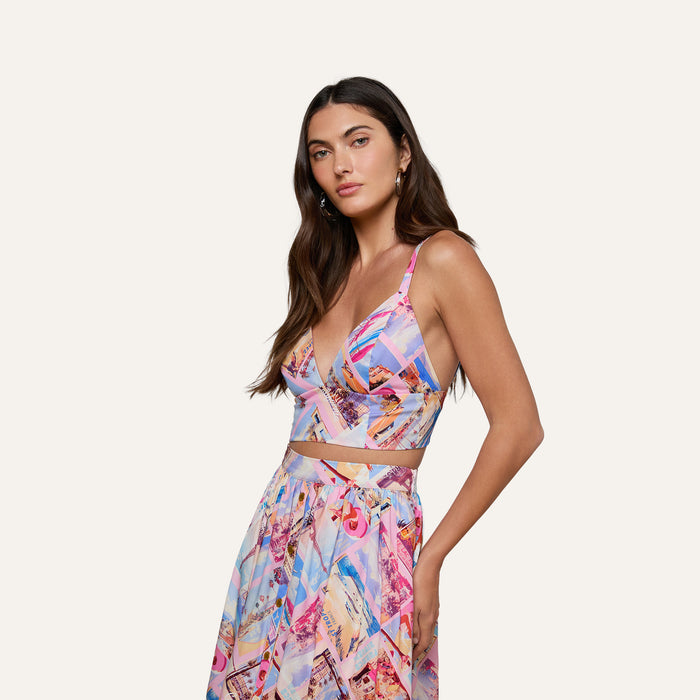 A woman with long, loosely styled dark hair stands confidently against a plain light background, wearing the Kaori Bralette Top in a colorful postcard print, paired with a matching skirt crafted from cotton-blend poplin.