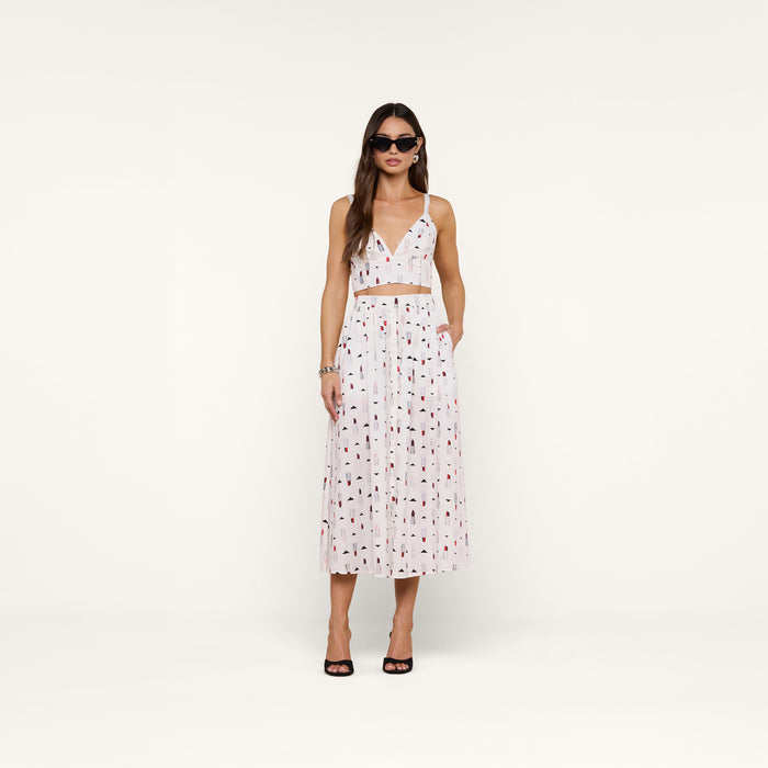 A woman wearing sunglasses and a white two-piece outfit featuring the Kaori Bralette Top with a lipstick print and a matching midi skirt, standing against a plain white background. She completes the look with black heeled sandals and gold jewelry.