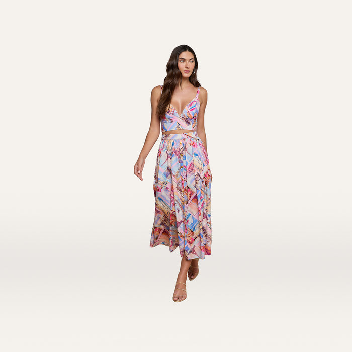A woman styled in the Kaori Bralette Top and a vibrant postcard print midi skirt, both crafted from cotton-blend poplin, paired with nude heels as she walks against a plain white background.