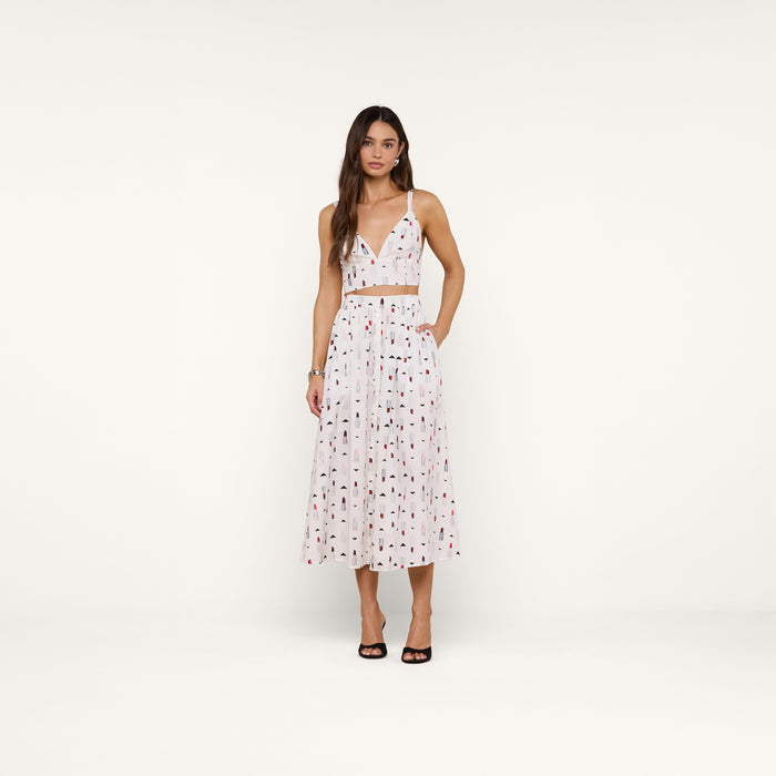 Woman wearing the Kaori Bralette Top styled with a white pleated midi skirt featuring small multicolored lipstick prints and a wide waist band, standing against a plain white background and pairing the look with black open-toe heels.
