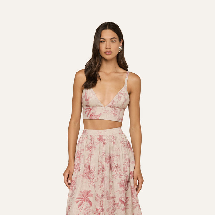 Wearing the Kaori Linen Bralette Top in light pink, a woman stands against a plain light background. She pairs the thin-strapped bralette with a matching high-waisted skirt featuring a palm tree pattern.