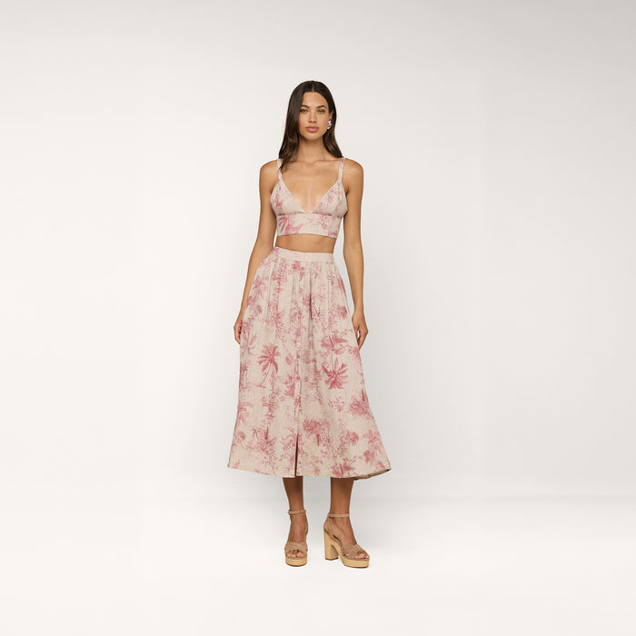 A woman stands in a studio wearing the light pink Kaori Linen Bralette Top in a jungle toile print, paired with a matching midi skirt and tan platform sandals.