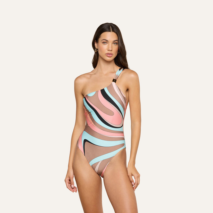 Wearing the Karmin One-Piece Swimsuit, which features a swirl print with a colorful abstract pattern in pink, brown, blue, and black, a woman stands against a plain light background.