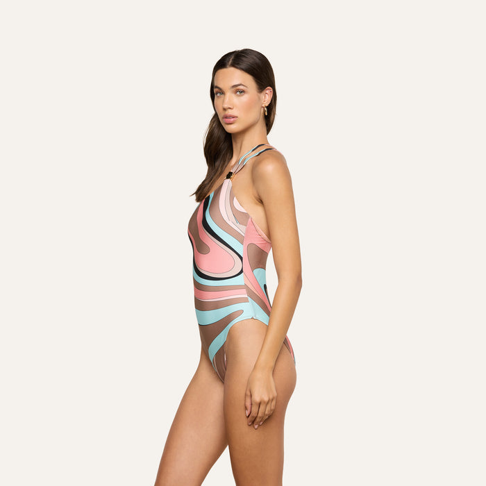 A woman stands sideways against a plain light background, looking at the camera with a neutral expression, wearing the Karmin One-Piece Swimsuit featuring a multicolored, patterned one-shoulder design.