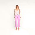 A woman stands against a plain background wearing a white tank top, the Kendra Coated Cropped Flare Jean in high-rise pink with a white belt, and white high-heeled sandals. She has blonde hair and a neutral expression.