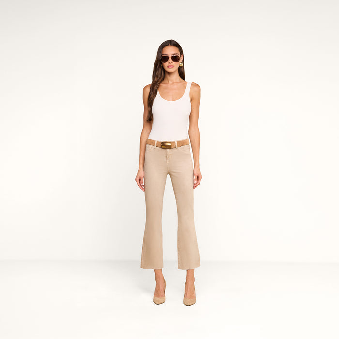 A woman stands against a plain white background, facing forward with her hands relaxed by her sides, wearing sunglasses, a white tank top, beige heels, and the Kendra Coated Cropped Flare Jean in high-rise beige.