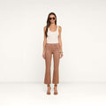A woman wearing sunglasses, a white tank top, the Kendra Coated Cropped Flare Jean in tan, and brown heeled sandals stands against a plain white background.
