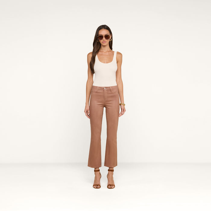 A woman wearing sunglasses, a white tank top, the Kendra Coated Cropped Flare Jean in tan, and brown heeled sandals stands against a plain white background.