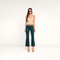 A woman stands against a plain white background wearing sunglasses, a beige sleeveless top, the Kendra Coated Cropped Flare Jean in dark green with a tan belt, and beige heels.