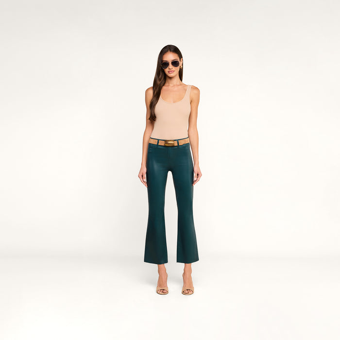 A woman stands against a plain white background wearing sunglasses, a beige sleeveless top, the Kendra Coated Cropped Flare Jean in dark green with a tan belt, and beige heels.