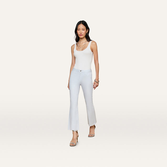 A woman wearing a white tank top, the Kendra Coated Cropped Flare Jean in a light hue, and heels stands against a plain, light background. She has long dark hair and accessorizes with layered necklaces and bracelets.