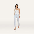 A woman wearing a white tank top, the Kendra Coated Cropped Flare Jean in a light hue, and heels stands against a plain, light background. She has long dark hair and accessorizes with layered necklaces and bracelets.