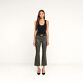 A woman stands against a plain white background, wearing the Kendra Coated Cropped Flare Jean in ash olive green, paired with a black tank top, black belt, and black heels. She has long brown hair and is looking directly at the camera.