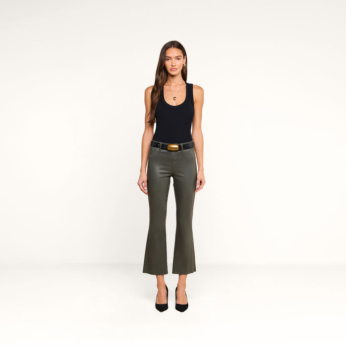 A woman stands against a plain white background, wearing the Kendra Coated Cropped Flare Jean in ash olive green, paired with a black tank top, black belt, and black heels. She has long brown hair and is looking directly at the camera.