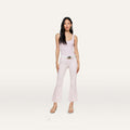 A woman with long dark hair and a neutral expression stands against a plain background, wearing a light pink sleeveless top paired with the Kendra Coated Cropped Flare Jean in a matching shade, styled with a white belt and beige heeled sandals.