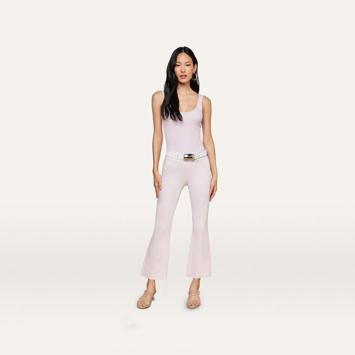 A woman with long dark hair and a neutral expression stands against a plain background, wearing a light pink sleeveless top paired with the Kendra Coated Cropped Flare Jean in a matching shade, styled with a white belt and beige heeled sandals.