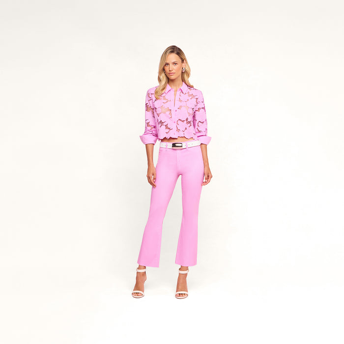 A woman stands against a white background wearing a pink floral blouse, the Kendra Coated Cropped Flare Jean in light pink high rise, a white belt, and nude heels.