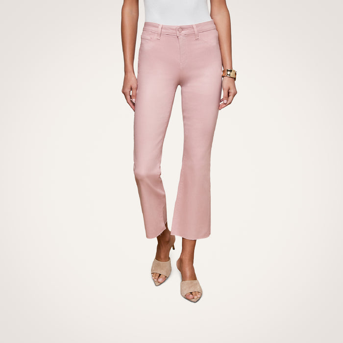 A woman wearing the Kendra Coated Cropped Flare Jean in blush pink, paired with a white sleeveless top, beige heeled sandals, and a gold watch, stands against a plain white background with only the lower half of her body visible.