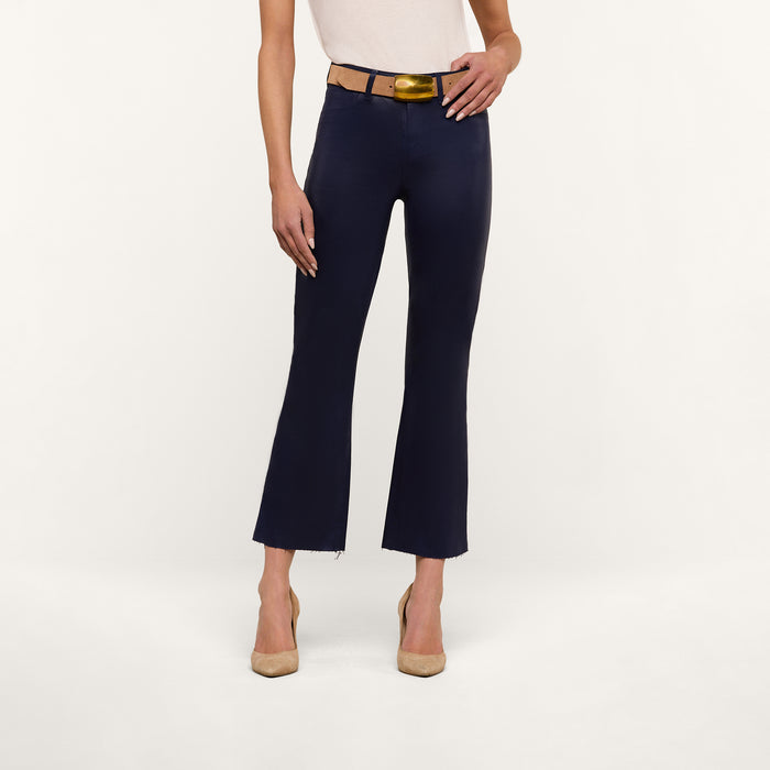 A person stands against a plain white background wearing a sleeveless beige top, a tan belt, beige heels, and the Kendra Coated Cropped Flare Jean in dark blue.