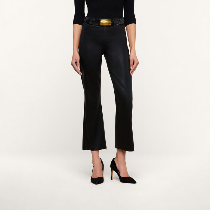 A woman is shown wearing the Kendra Coated Cropped Flare Jean in black, styled with a matching long-sleeve top, high heels, and a statement belt with a large gold buckle. She stands against a plain white background, with her head and feet cropped out of the image.