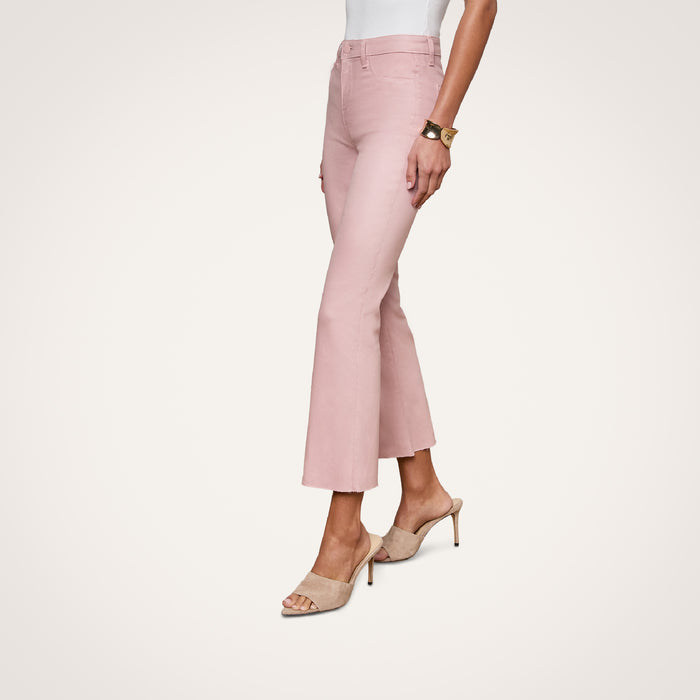 A woman wearing the Kendra Coated Cropped Flare Jean in pink, paired with a white top, beige heeled sandals, and a bracelet, stands against a plain white background.