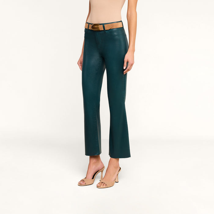A woman wearing the Kendra Coated Cropped Flare Jean in teal, paired with a tan belt, a sleeveless beige top, and nude high-heeled sandals stands against a white background.