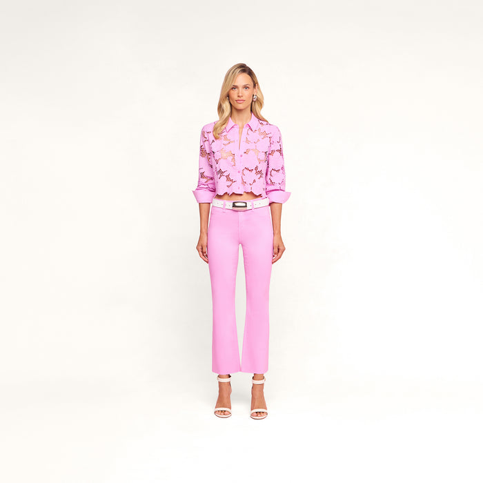 A woman with blonde hair and a neutral expression stands against a white background wearing the Kendra Coated Cropped Flare Jean in pink, paired with a light pink floral blouse, a white belt, and white heeled sandals.