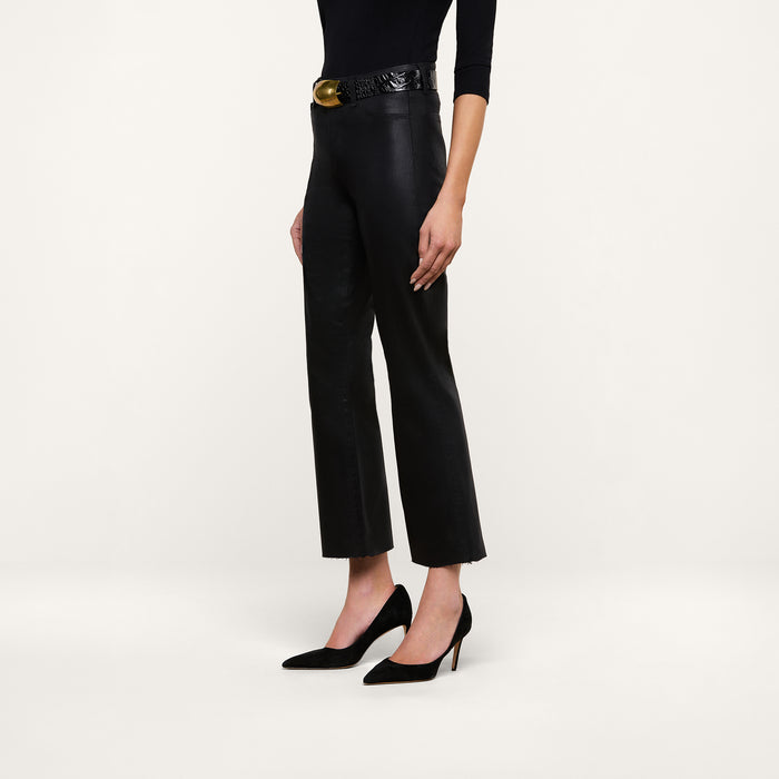 A person stands against a plain white background, wearing the Kendra Coated Cropped Flare Jean with a high-rise fit, styled with a black belt featuring a gold buckle, a long-sleeve black top, and black high-heeled shoes.