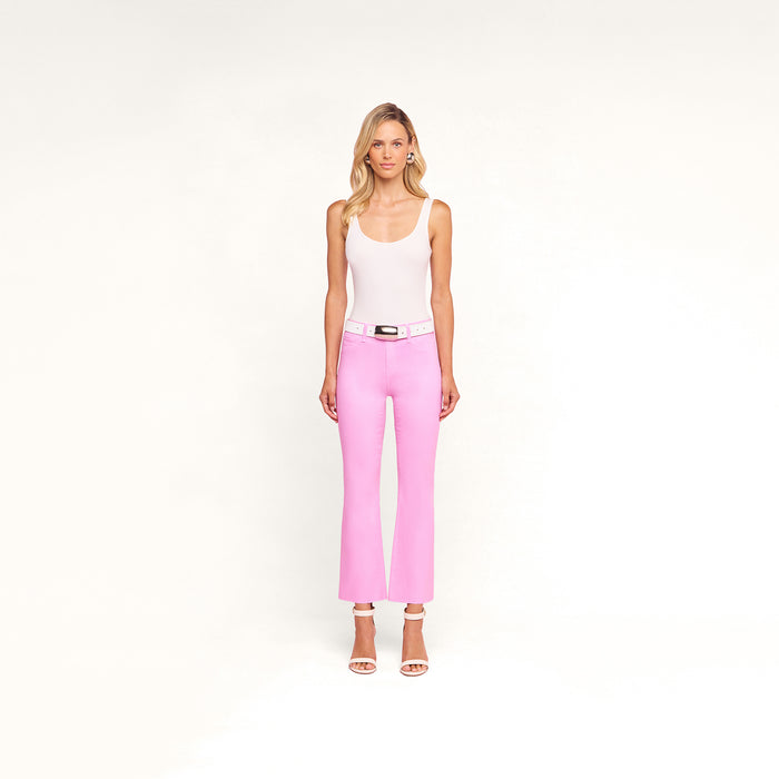A woman stands facing forward against a plain white background, wearing the Kendra Coated Cropped Flare Jean in pink with a white belt, paired with a white tank top and white high-heeled sandals.