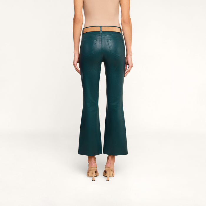 A person stands facing away, wearing the Kendra Coated Cropped Flare Jean in dark teal, paired with a tan sleeveless top and tan high-heeled shoes against a plain white background.
