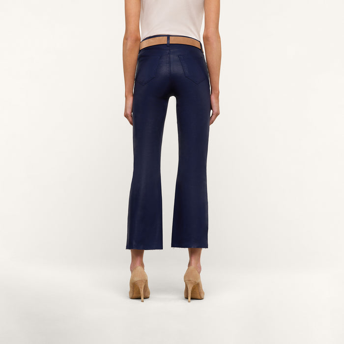 A person wearing the Kendra Coated Cropped Flare Jean in dark blue, paired with a beige top and tan high-heeled shoes, is standing facing away from the camera against a plain white background.