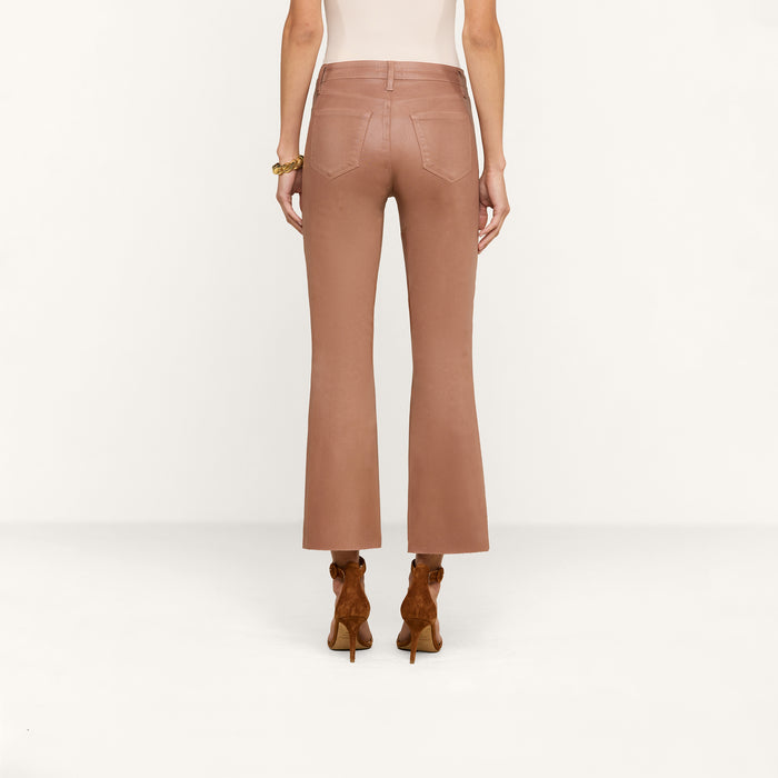 A woman stands facing away against a plain white background, hands relaxed at her sides, wearing the Kendra Coated Cropped Flare Jean in light brown paired with brown heeled shoes.