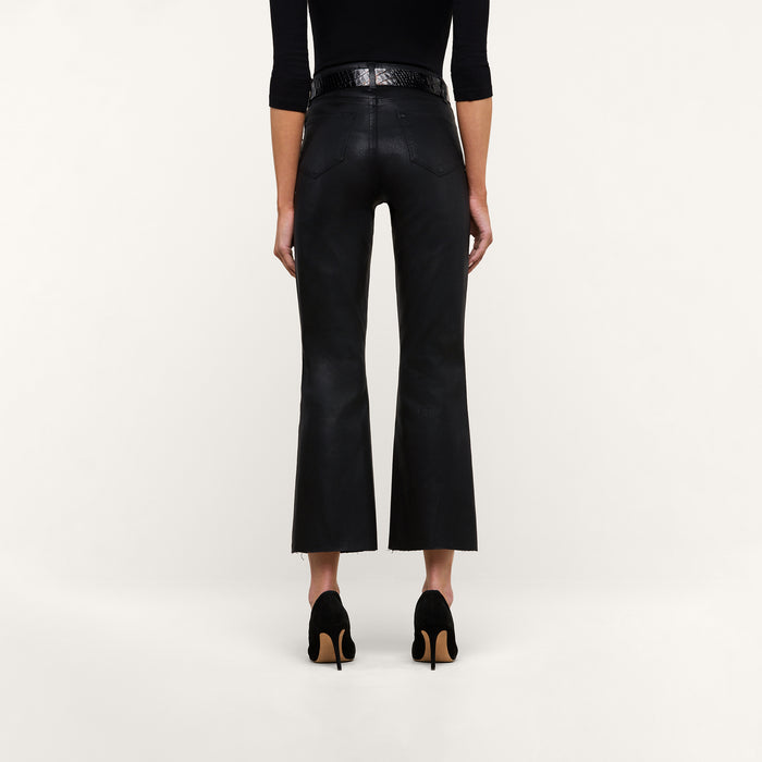 A person stands facing away against a plain white background, wearing the Kendra Coated Cropped Flare Jean in high-rise black coated denim, styled with a black belt, a black long-sleeve top, and black heeled shoes.