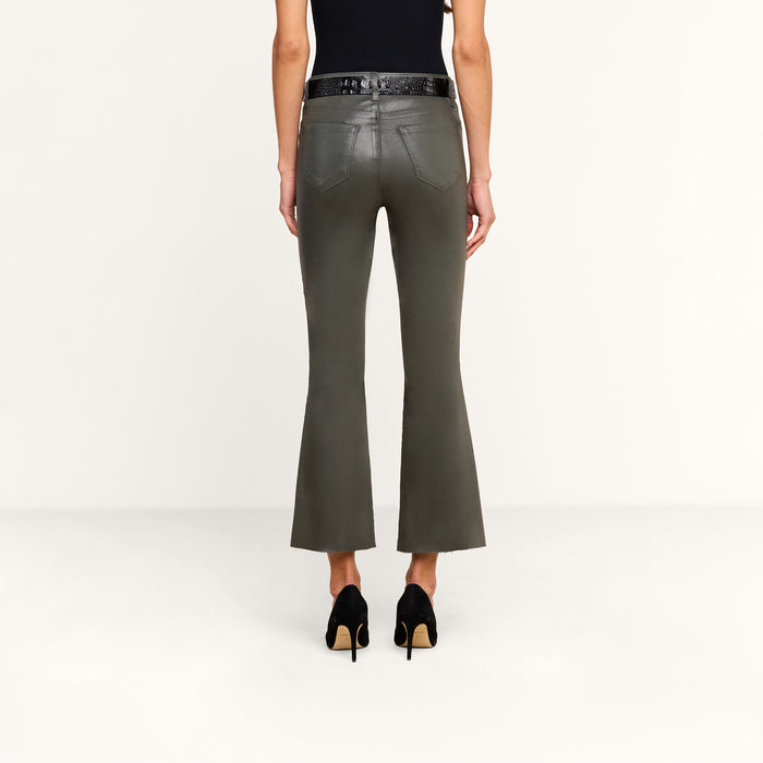A person stands facing away against a plain white background, wearing the Kendra Coated Cropped Flare Jean in ash olive green, paired with a black belt, a black top, and black high-heeled shoes.