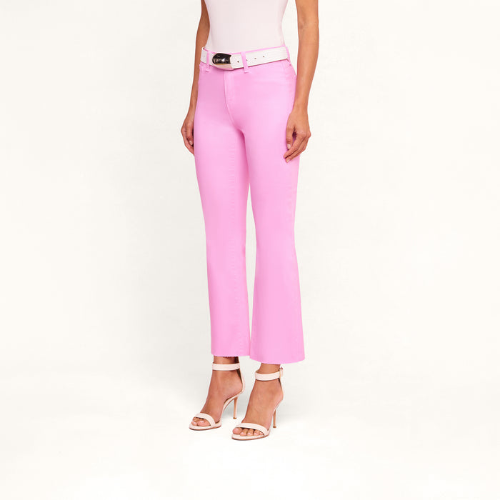 A woman stands against a plain white background wearing the Kendra Coated Cropped Flare Jean in light pink with a white belt, a white sleeveless top, and white high-heeled sandals. Her head is not visible in the image.