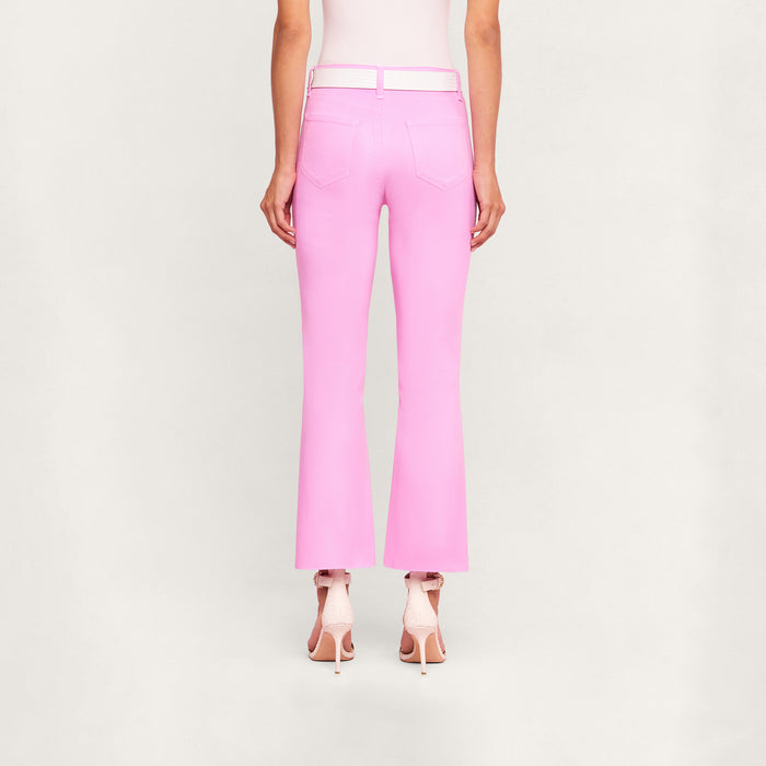A person stands facing away, wearing the Kendra Coated Cropped Flare Jean in light pink crafted from coated denim, paired with high-heeled pink shoes against a plain, light background.