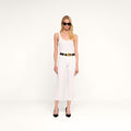 A woman with long blonde hair, wearing a white sleeveless top, the Kendra Cropped Flare Jean in white stretch denim with a raw hem, a black belt with a gold buckle, black high heels, and black sunglasses, stands against a plain white background.