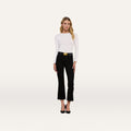 A woman stands facing forward, wearing a white long-sleeve top and the Kendra Cropped Flare Jean—premium high-rise cropped flare jeans in a black hue with frayed hems—paired with a black belt featuring a gold buckle and black heels, against a plain white background.