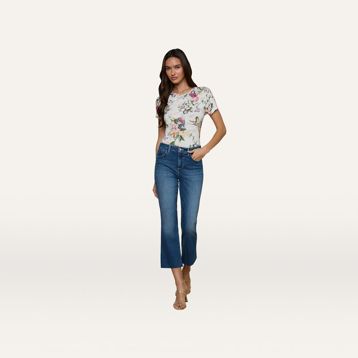 A woman stands against a plain white background, wearing a floral short-sleeve top, beige heeled sandals, and the Kendra Cropped Flare Jean—a pair of blue high-waisted cropped flare jeans in a dark denim wash. She has long brown hair and one hand in her pocket.